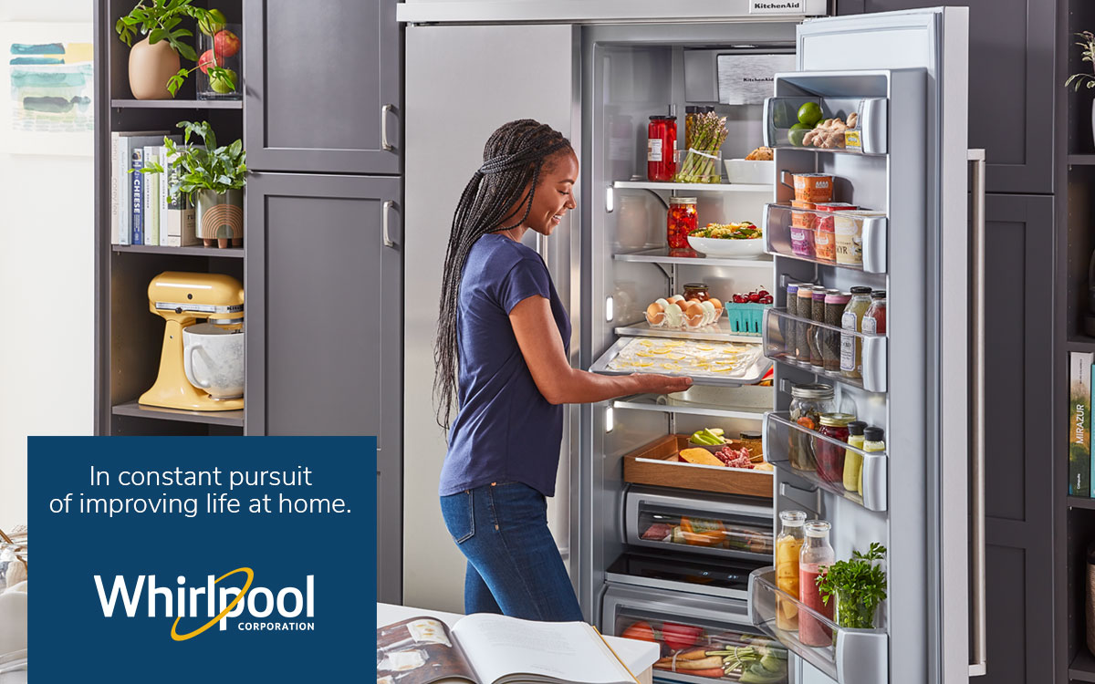 Smiling Black woman in kitchen, placing a sheetcake topped with whipped cream and lemon slices and zest in her refrigerator. Cookbook and baking ingredients are in foreground on counter.